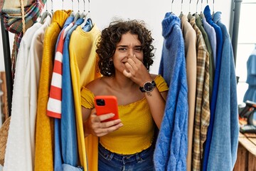 Young hispanic woman searching clothes on clothing rack using smartphone smelling something stinky and disgusting, intolerable smell, holding breath with fingers on nose. bad smell