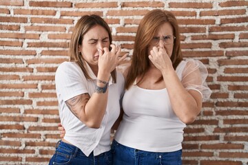 Hispanic mother and daughter wearing casual white t shirt smelling something stinky and disgusting, intolerable smell, holding breath with fingers on nose. bad smell - obrazy, fototapety, plakaty