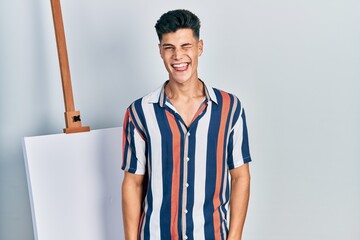 Young hispanic man standing close to empty canvas winking looking at the camera with sexy...