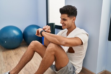 Young hispanic man sitting on floor using watch at sport center