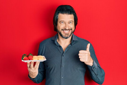 Middle age caucasian man eating fresh and healthy fruit smiling happy and positive, thumb up doing excellent and approval sign