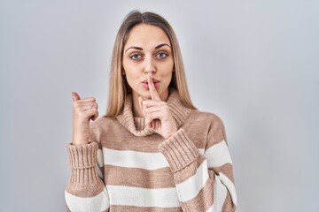 Young blonde woman wearing turtleneck sweater over isolated background asking to be quiet with finger on lips pointing with hand to the side. silence and secret concept.