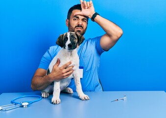 Handsome hispanic veterinary man with beard checking dog health making fun of people with fingers...