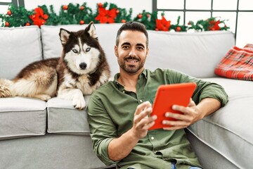 Young hispanic man using touchpad sitting on sofa with dog by christmas decor at home