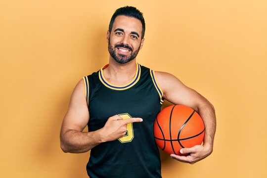 Handsome hispanic man with beard holding basketball ball smiling happy pointing with hand and finger