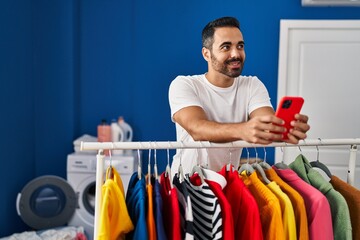 Young hispanic man using smartphone leaning on clothes rack at laundry room