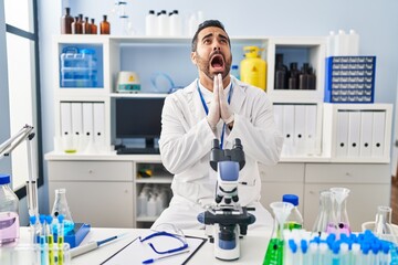 Young hispanic man with beard working at scientist laboratory begging and praying with hands...