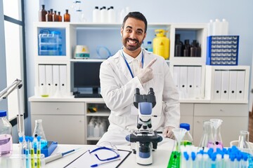 Young hispanic man with beard working at scientist laboratory cheerful with a smile of face pointing with hand and finger up to the side with happy and natural expression on face