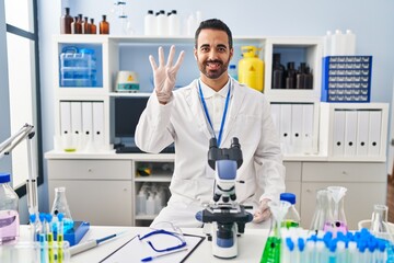 Young hispanic man with beard working at scientist laboratory showing and pointing up with fingers number four while smiling confident and happy.