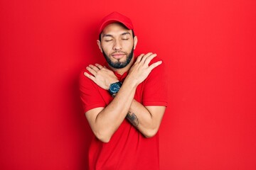 Hispanic man with beard wearing delivery uniform and cap hugging oneself happy and positive, smiling confident. self love and self care