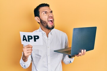 Hispanic man with beard holding computer laptop and app banner angry and mad screaming frustrated and furious, shouting with anger looking up.