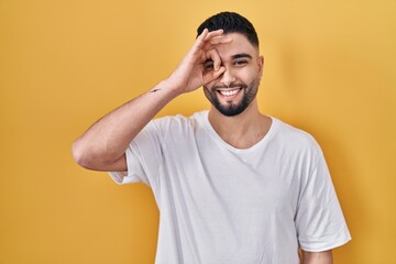 Fototapeta na wymiar Young handsome man wearing casual t shirt over yellow background doing ok gesture with hand smiling, eye looking through fingers with happy face.