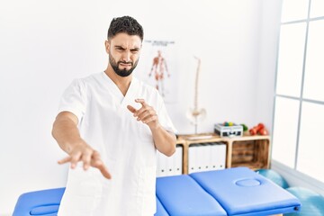 Young handsome man with beard working at pain recovery clinic disgusted expression, displeased and fearful doing disgust face because aversion reaction. with hands raised