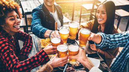 Group of multiracial friends having bbq dinner party together - Diverse young people sitting at bar table toasting beer glasses in brewery pub garden - Brewery, dining lifestyle and food concept