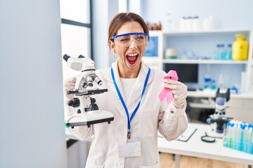 Young brunette woman working at scientist laboratory holding pink ribbon winking looking at the camera with sexy expression, cheerful and happy face.
