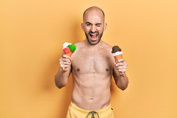 Young bald man wearing swimwear holding two ice cream cones winking looking at the camera with sexy...