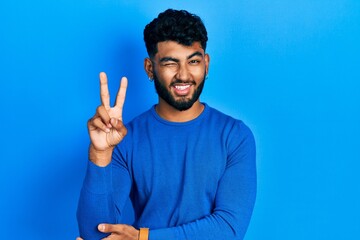 Arab man with beard wearing casual blue sweater smiling with happy face winking at the camera doing victory sign. number two.