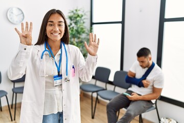 Young asian doctor woman at waiting room with a man with a broken arm showing and pointing up with fingers number nine while smiling confident and happy.
