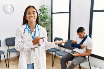Fototapeta premium Young asian doctor woman at waiting room with a man with a broken arm amazed and smiling to the camera while presenting with hand and pointing with finger.