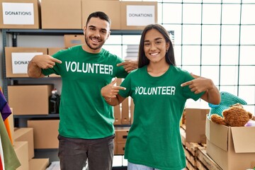 Young latin couple smiling happy pointing to volunteer uniform at charity center.