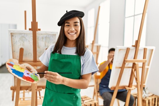 Young hispanic couple drawing at art studio. Woman smiling happy and holding paintbrush and palette.