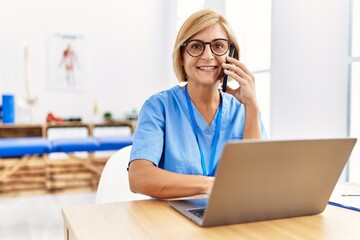 Middle age blonde woman wearing physio therapy uniform talking on the smartphone and using laptop at clinic - Powered by Adobe