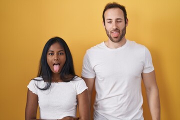 Obraz premium Interracial couple standing over yellow background sticking tongue out happy with funny expression. emotion concept.