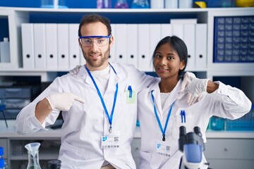 Interracial couple working at scientist laboratory pointing finger to one self smiling happy and proud