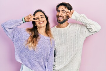 Young hispanic couple wearing casual clothes doing peace symbol with fingers over face, smiling cheerful showing victory