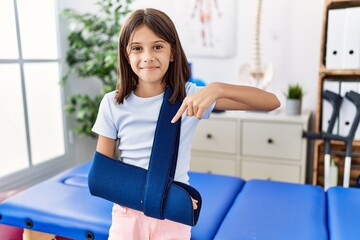 Young hispanic girl wearing arm on sling at rehabilitation clinic smiling happy pointing with hand...