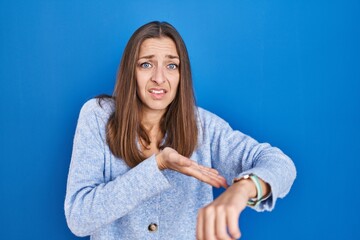 Young woman standing over blue background in hurry pointing to watch time, impatience, upset and...