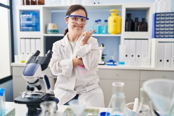 Hispanic girl with down syndrome working at scientist laboratory with a big smile on face, pointing with hand finger to the side looking at the camera.
