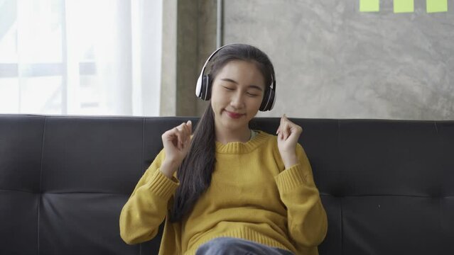 Asian woman wearing headphones listening to music happily on a sopa at home Video 4k