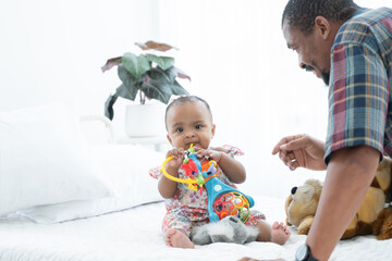 African adorable newborn baby daughter playing and bite toy in her hands while sitting on bed with father taking care of little child at home. Baby teething concept.