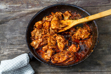 Pieces of baked chicken in sauce in a frying pan with a wooden spatula on a dark table.