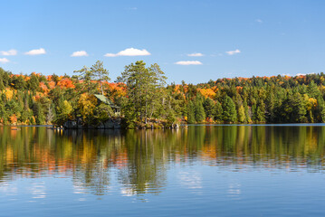 Cabin perched on a small rocky island in the middle of a lake surrounded by forest on a sunny...