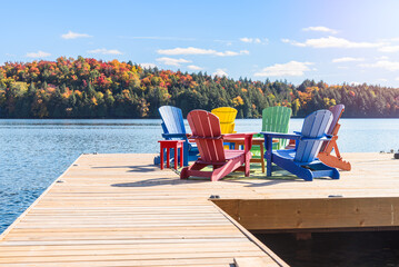 Colourful adirondack chairs around atable on a wooden pier on a lake on a sunny autumn day.