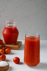A glass of tomato juice, next to a jug of juice, tomatoes and salt.