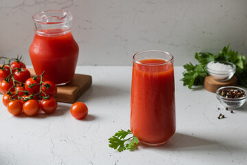 A glass of tomato juice, next to a jug of juice, tomatoes, parsley and pepper and salt.