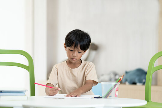 Young asian child boy using pencil writing something in book at classroom. Asian son doing homework while sitting at desk at home. Kid learning by drawing. Education concept
