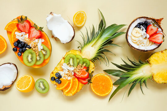 Healthy fruit yogurt bowls in a pineapple, papaya and coconut. Overhead view table scene on a pale yellow background.