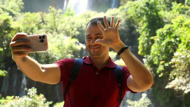 Smiling adult man blogger, vlogger recording selfie movie on smartphone at nature, waterfall background. Happy social media content maker, influencer show thumbs up, victory sign hand on phone camera.