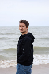 A young man in a black hoodie on the beach against the backdrop of the sea. Brown hair, blue eyes, smiling