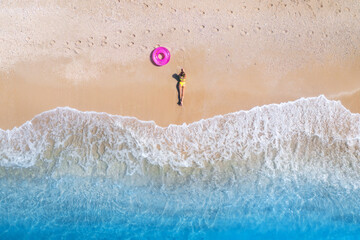 Fototapeta Aerial view of the lying beautiful young woman with pink swim ring on the sandy beach near sea with waves at sunset. Summer vacation in Lefkada island, Greece. Top view of sexy girl, clear blue water obraz