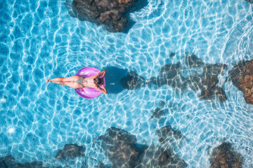 Aerial view of a young woman swimming with pink swim ring in blue sea at sunset in summer. Tropical landscape with sexy girl, clear water, stones, sandy beach. Top view. Lefkada island, Greece. Nature