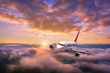 Naklejka na ściany i meble Airplane is flying above the clouds at sunset in summer. Landscape with passenger airplane, mountains, orange sky. Aircraft is taking off. Business travel. Commercial plane. Aerial view. Transport 