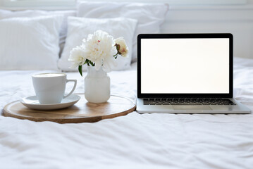 Light cozy bedroom, mock up laptop, coffee cup and an flowers on the white bed. Work from bedroom. Coffee cup and flowers on a white bed. White Concept. Remote work, empty screen. White screen. 