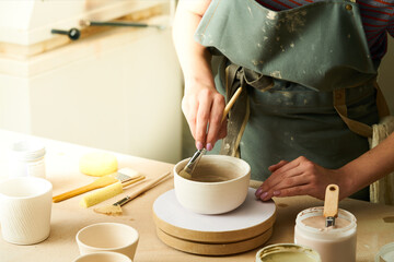 Close-up of girl painting clay mug with glaze. Woman coloring pottery in workshop with a paintbrush. Painter in green apron glazing clay pot.