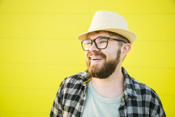 Portrait of positive young hipster man smiling over yellow wall store outdoor background - Handsome trendy bearded guy with hat and glasses standing outdoors - Fashion and trendy youth people concept