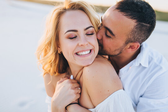 A tanned young caucasian bearded guy kisses a beautiful fun smiling blonde woman against the backdrop of the setting sun. Desert, sandy beach, rest and relaxation. Concept of honeymoon.